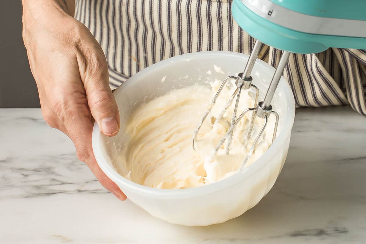 A woman mixing vanilla frosting with an electric hand mixer.