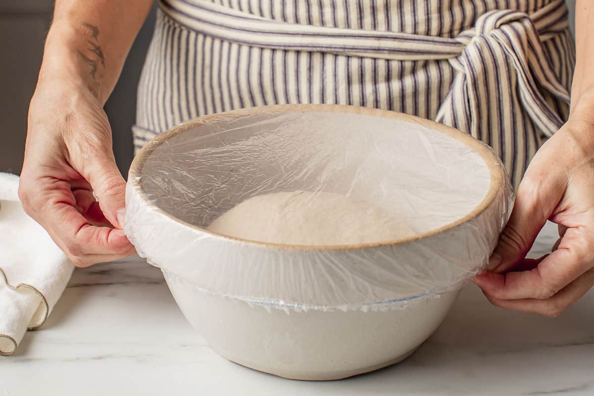 A woman covering a bowl of sourdough breadstick dough with plastic.