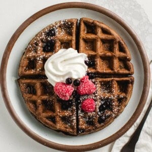 Chocolate sourdough waffles on a plate with berries and whipped cream and a fork.