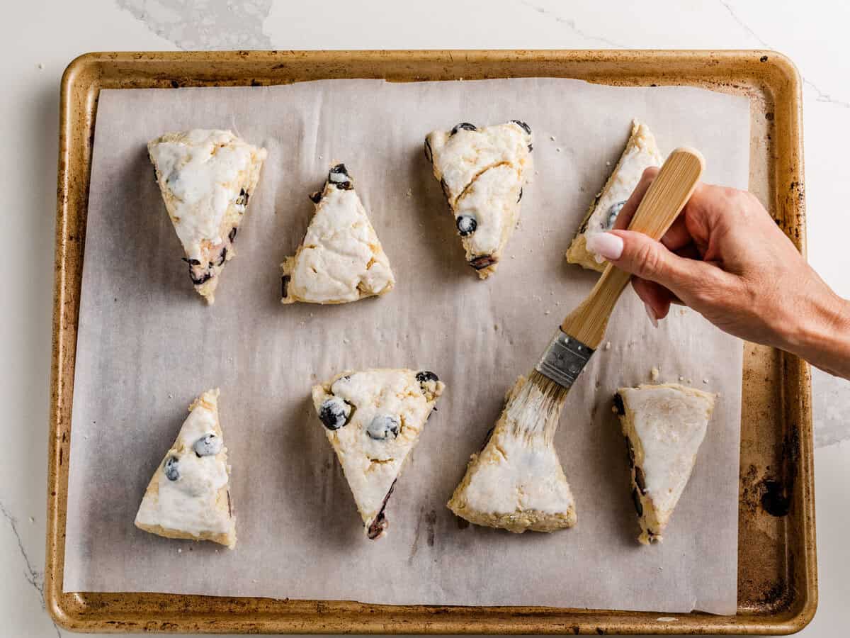 A woman brushing heavy cream onto each sourdough lemon blueberry scone on a baking sheet.