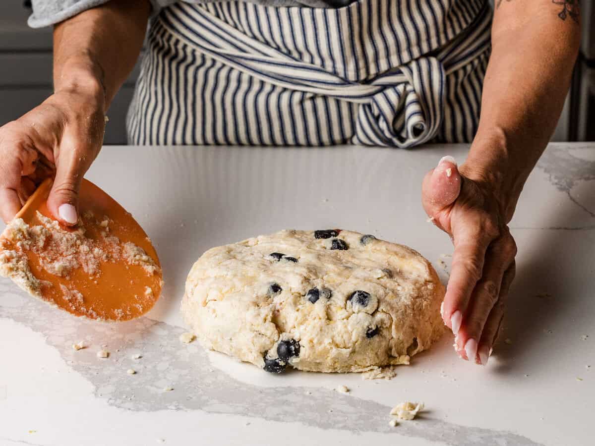 A woman forming scone dough into a disk on a table using her hands.