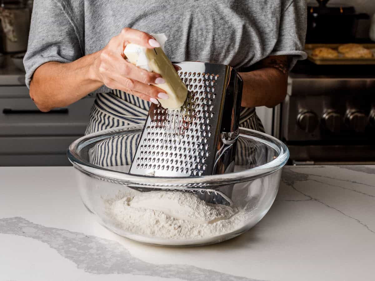 A woman grating butter into a bowl of flour, salt, and baking powder.