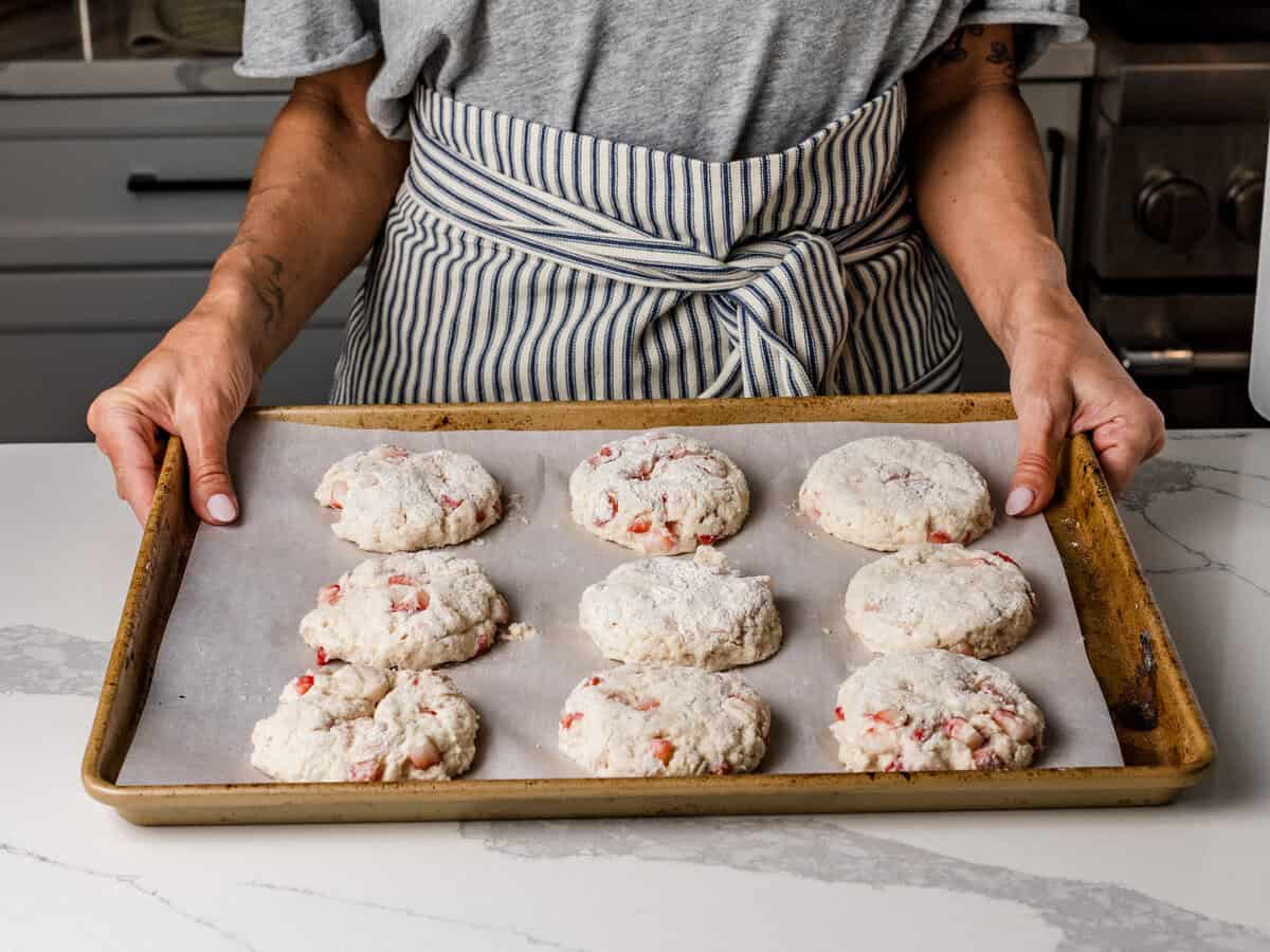 A woman holding a baking sheet lined with unbaked sourdough strawberry biscuits.