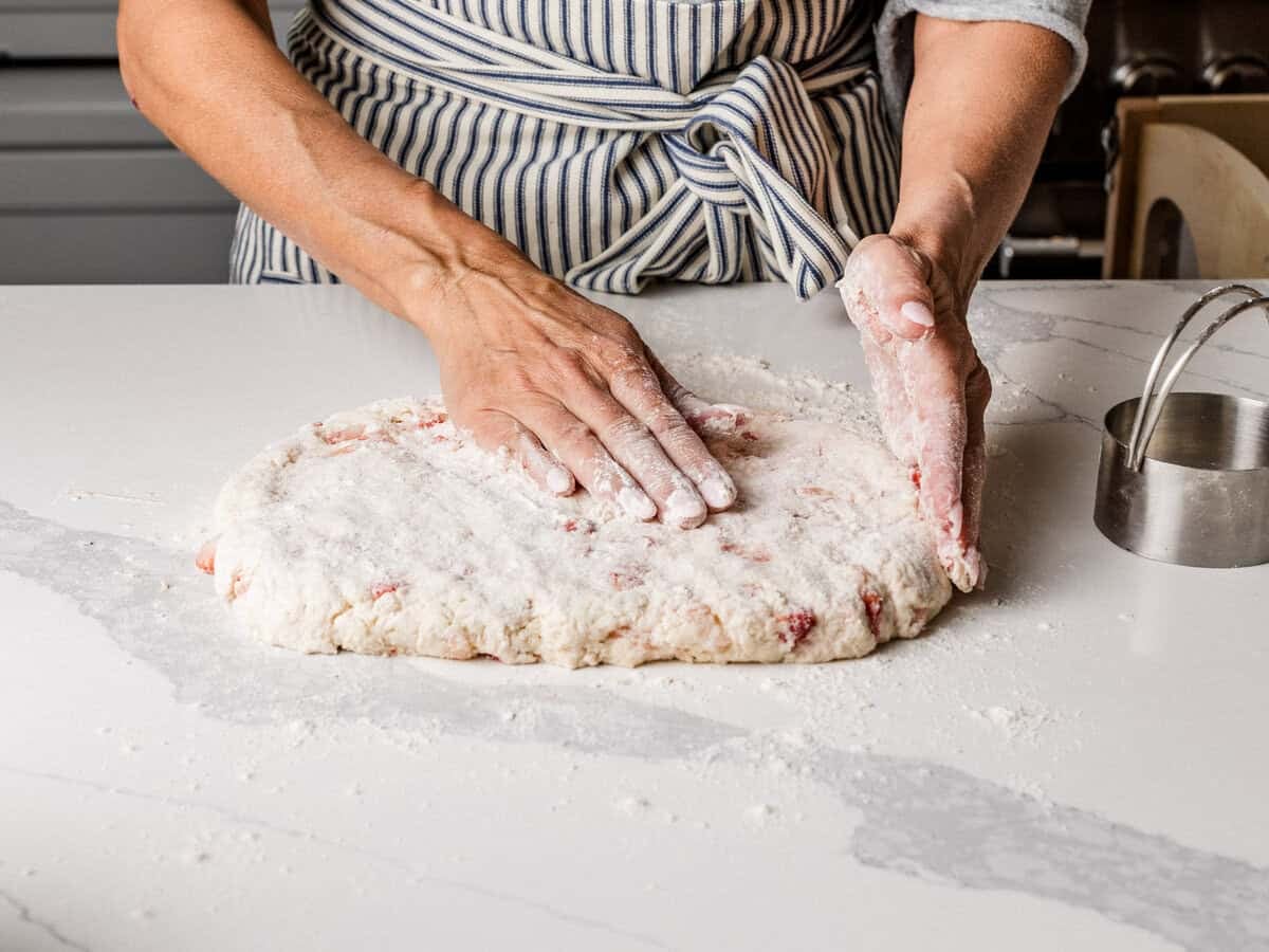 A woman using your hands to press out sourdough strawberry biscuit dough on a floured surfaced table.